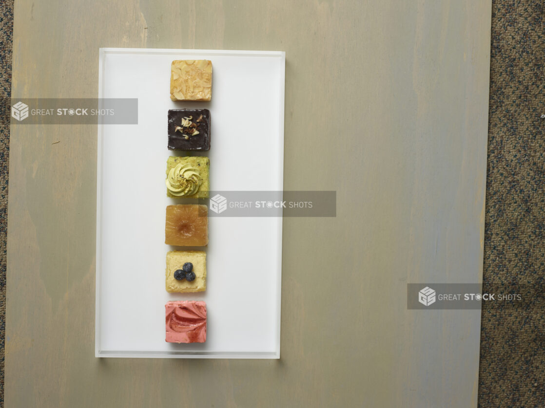 Elegant assorted mini square colourful desserts on a white rectangular platter on a wooden background