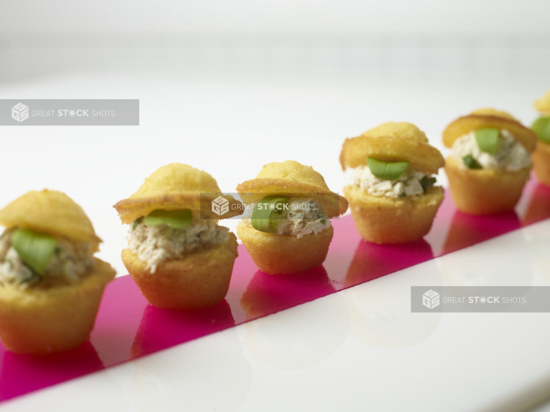 A Row of Mini Cornmeal Muffins Stuffed with Tuna Salad and Micro Greens on a Pink Plexiglass Strip on a White Surface