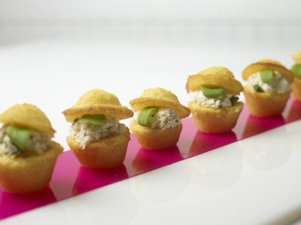 A Row of Mini Cornmeal Muffins Stuffed with Tuna Salad and Micro Greens on a Pink Plexiglass Strip on a White Surface