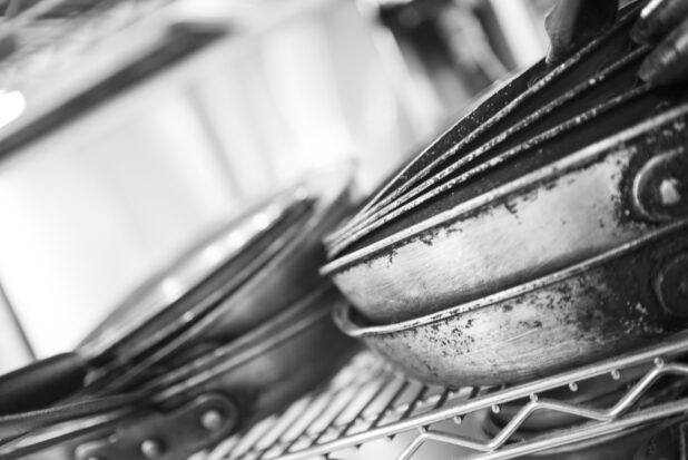 Black and White Photo of a Close Up of Well Worn Kitchen Sauce Pans Stacked on a Metal Wire Rack in a Restaurant Kitchen Setting