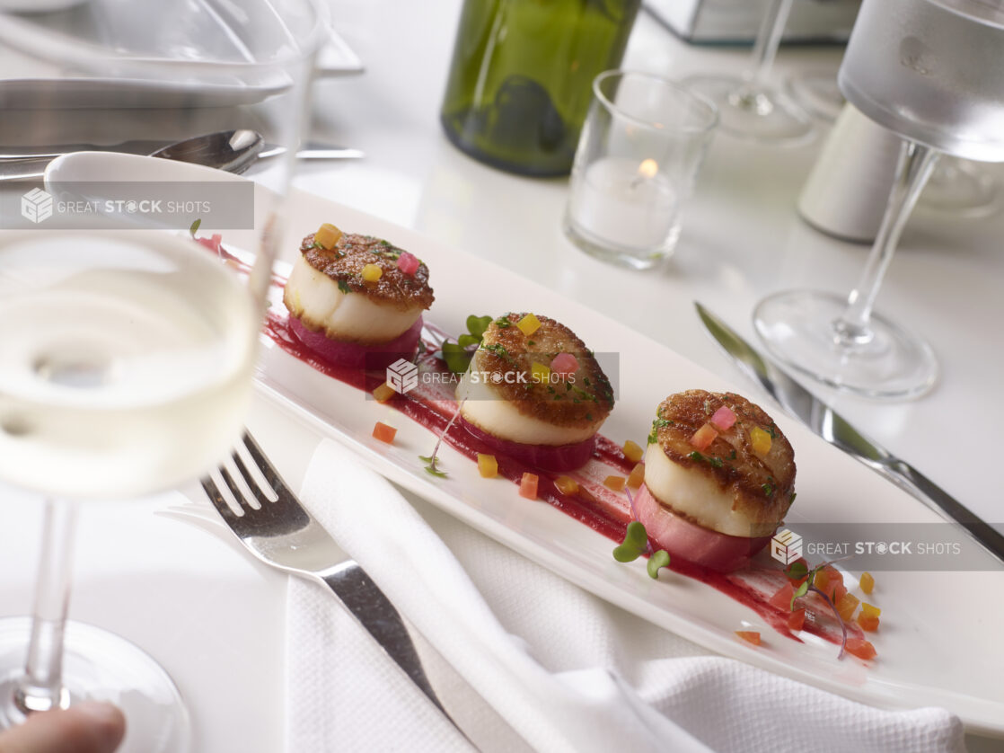 Pan seared scallops on poached radish on a long oval platter in an elegant table setting on white linen