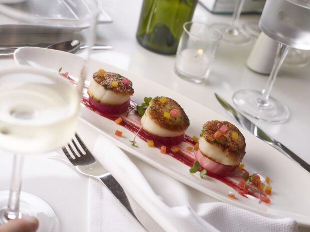 Pan seared scallops on poached radish on a long oval platter in an elegant table setting on white linen