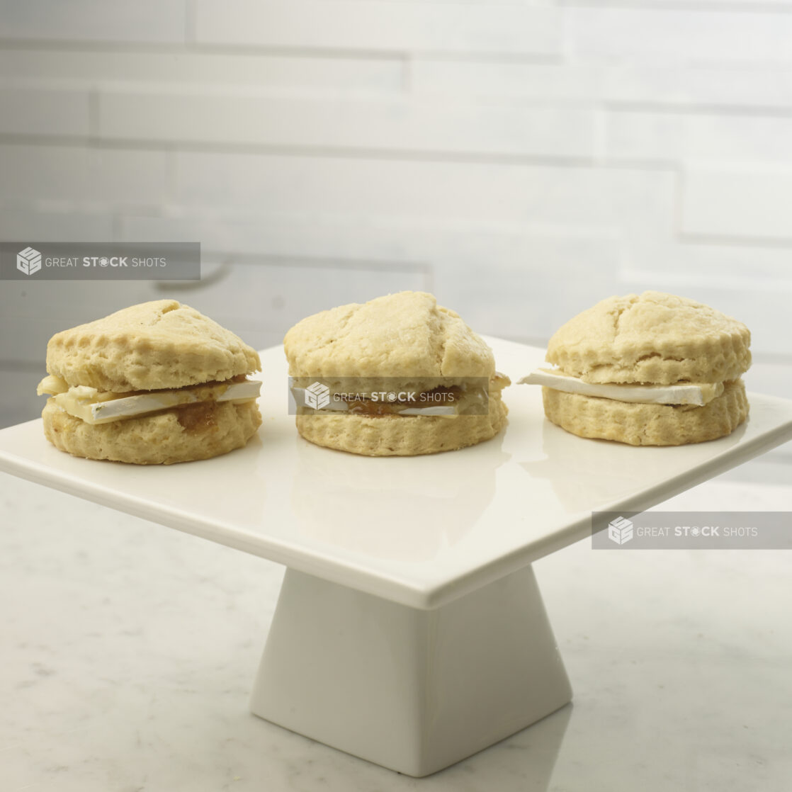 Tea biscuits with brie cheese and jam on a white ceramic cake stand on a white marble background