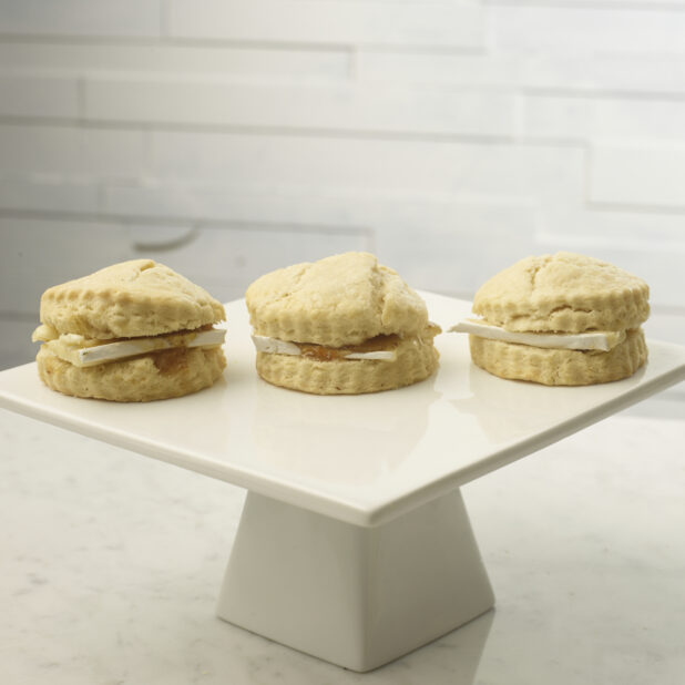 Tea biscuits with brie cheese and jam on a white ceramic cake stand on a white marble background