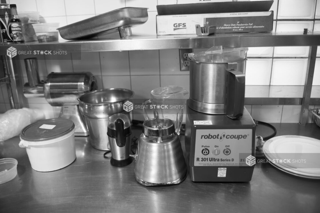 Black and white photo of a food processor, blender, coffee grinder, meat grinder on a stainless steel table in a restaurant kitchen