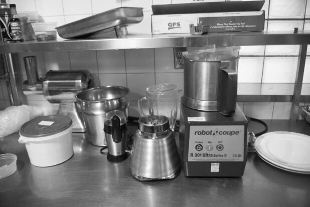 Black and white photo of a food processor, blender, coffee grinder, meat grinder on a stainless steel table in a restaurant kitchen