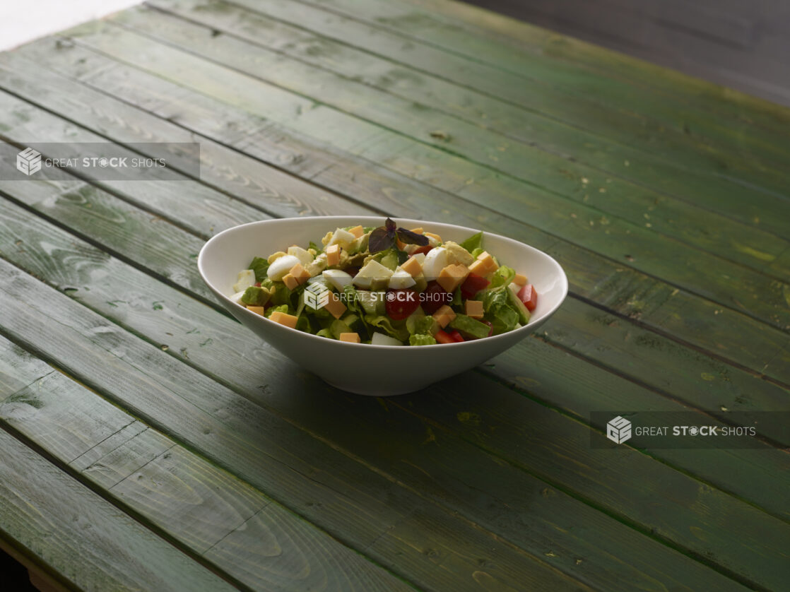 Green salad with red bell pepper, cheese, tomatoes and boiled egg in a white bowl on a hunter green wooden background