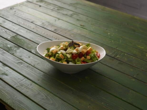Green salad with red bell pepper, cheese, tomatoes and boiled egg in a white bowl on a hunter green wooden background
