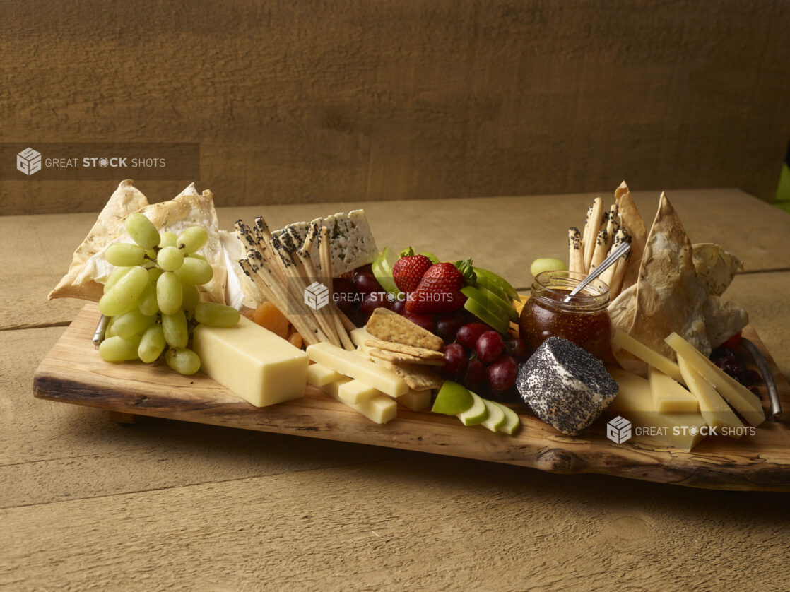 Rustic cheese tray with various cheese, bread sticks, flatbreads, fresh fruit and fig jam in a jar on a rustic wooden serving board