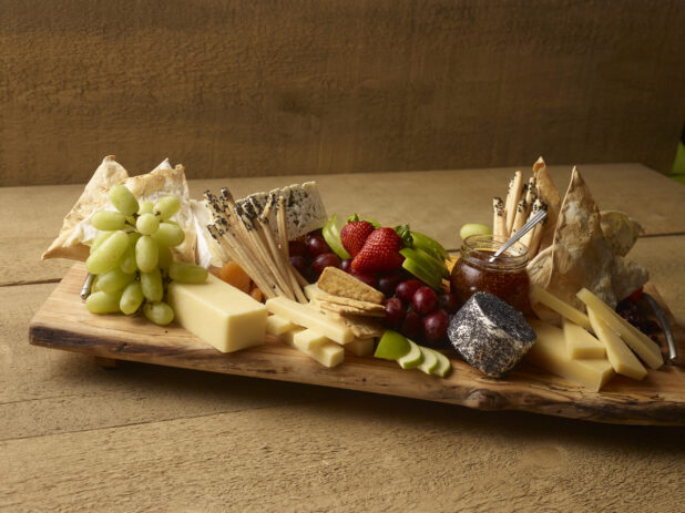 Rustic cheese tray with various cheese, bread sticks, flatbreads, fresh fruit and fig jam in a jar on a rustic wooden serving board