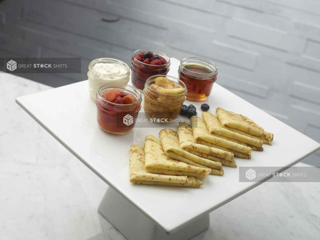 Crepes on a white ceramic cake stand with mason jars of mixed berries, honey, sauteed apples, cooked rhubarb and sour cream