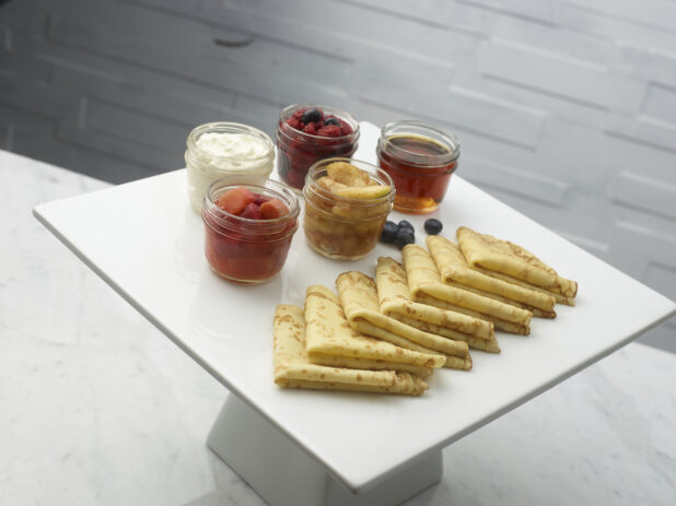 Crepes on a white ceramic cake stand with mason jars of mixed berries, honey, sauteed apples, cooked rhubarb and sour cream