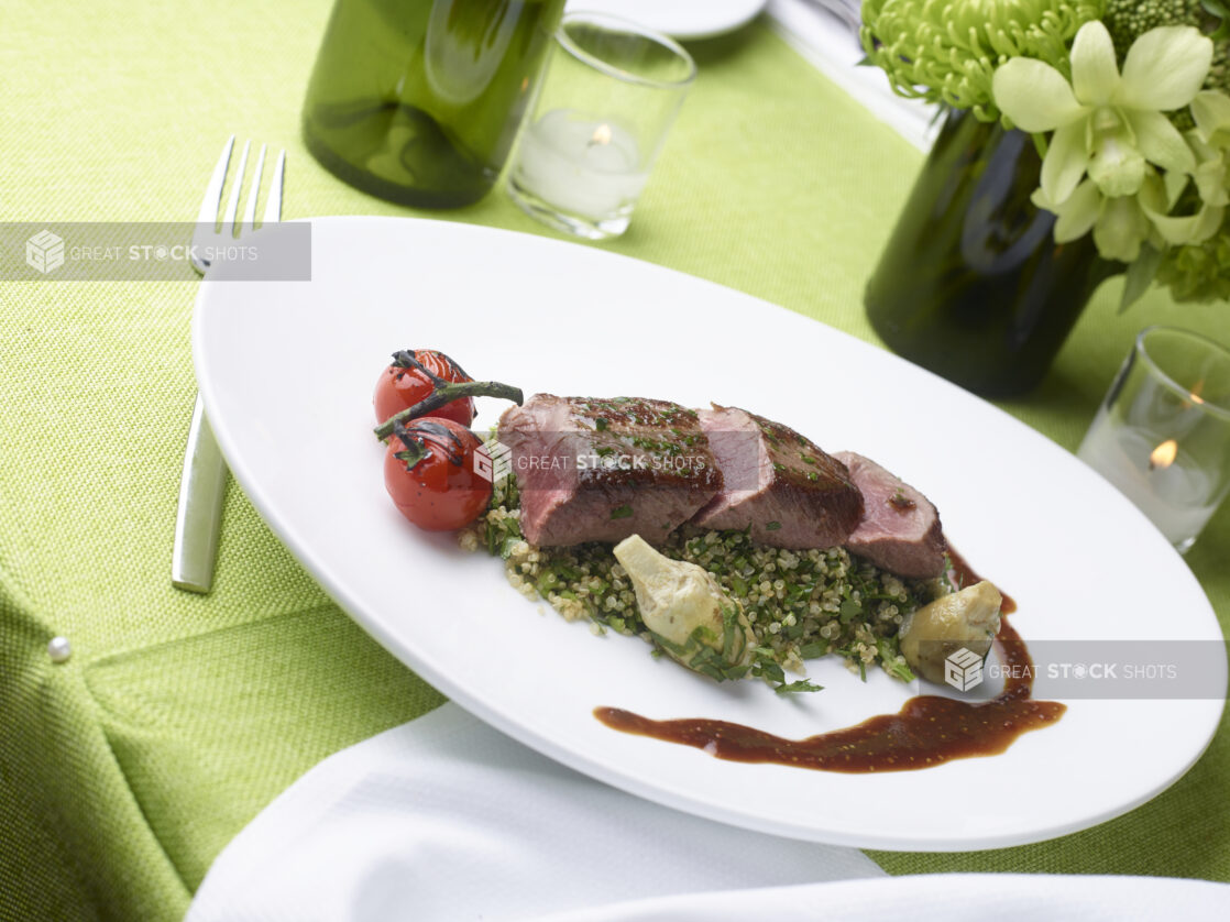 Grilled pork tenderloin over quinoa salad with grilled cherry tomato and artichoke hearts on a lime green tablecloth, elegant setting