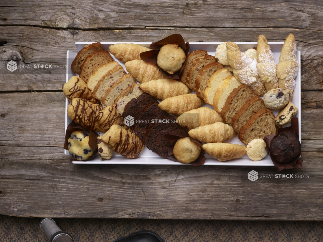 Assorted pastries, chocolate danish, muffins, croissants, almond croissant, loaves, breakfast bites on a white rectangular tray on a weathered wooden board