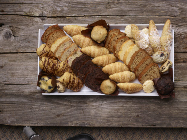Assorted pastries, chocolate danish, muffins, croissants, almond croissant, loaves, breakfast bites on a white rectangular tray on a weathered wooden board