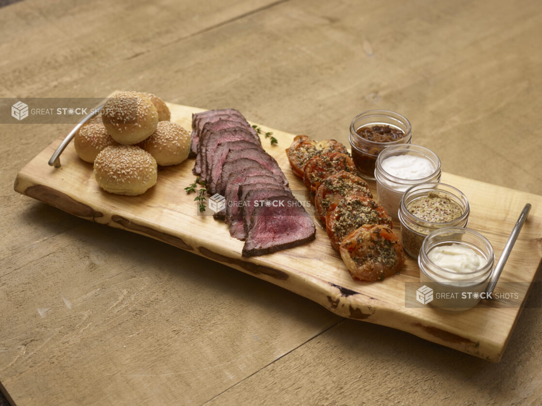 Rare sliced beef tenderloin with roasted tomatoes, mini sesame buns with mayo, aioli, grainy mustard and caramelized onions in mason jars on a wooden serving tray