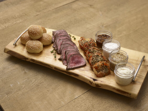 Rare sliced beef tenderloin with roasted tomatoes, mini sesame buns with mayo, aioli, grainy mustard and caramelized onions in mason jars on a wooden serving tray