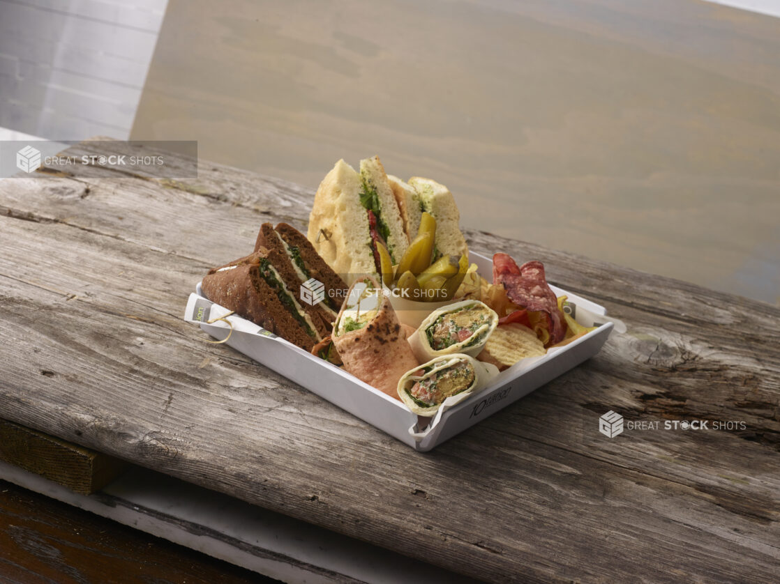 Artisanal sandwiches and wraps in a white square tray with potato chips and pickles on a weathered wooden board