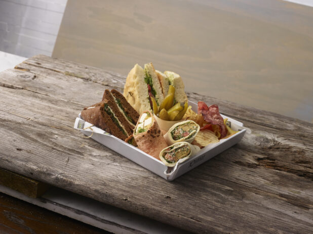 Artisanal sandwiches and wraps in a white square tray with potato chips and pickles on a weathered wooden board