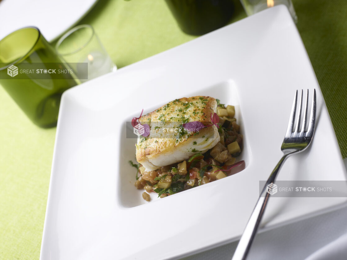 Pan seared cod over a homemade relish in a white square unique bowl with a fork resting on the bowl on a lime green background