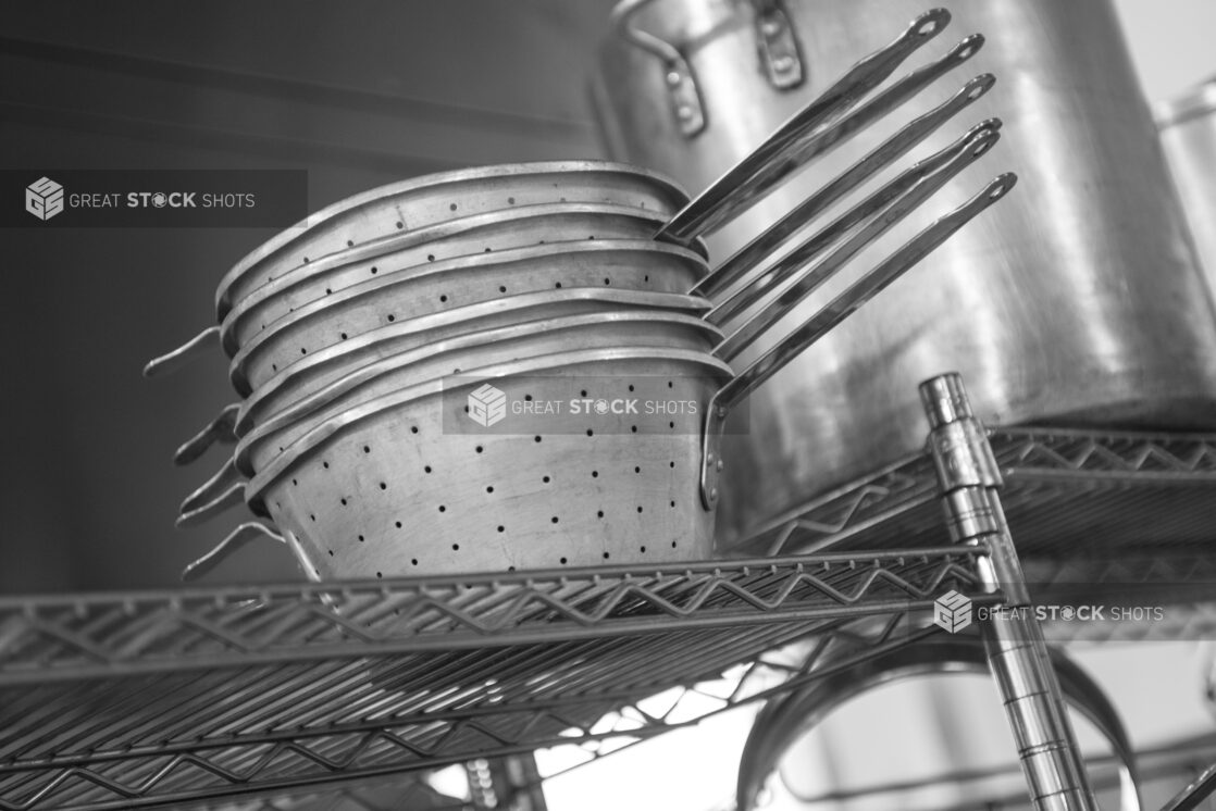 Black and white photo of metal strainers and stock pot on wire rack in a restaurant kitchen