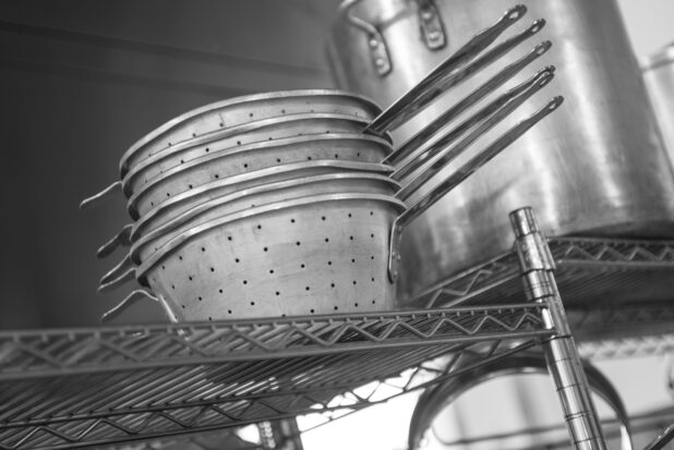 Black and white photo of metal strainers and stock pot on wire rack in a restaurant kitchen