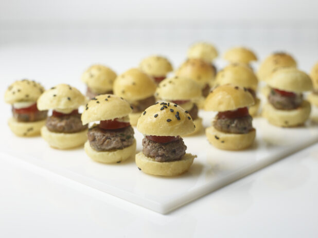 Mini homemade hamburger sliders on black sesame seed buns topped with sliced tomato and cheese on a rectangular white board