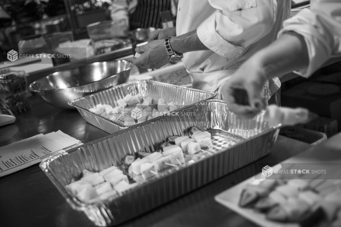 Black and white photo of chefs preparing fruit kabobs in a restaurant kitchen using disposable aluminum hotel pans