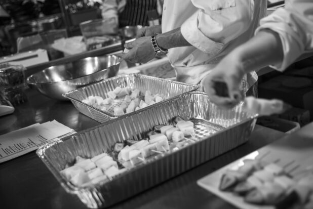 Black and white photo of chefs preparing fruit kabobs in a restaurant kitchen using disposable aluminum hotel pans