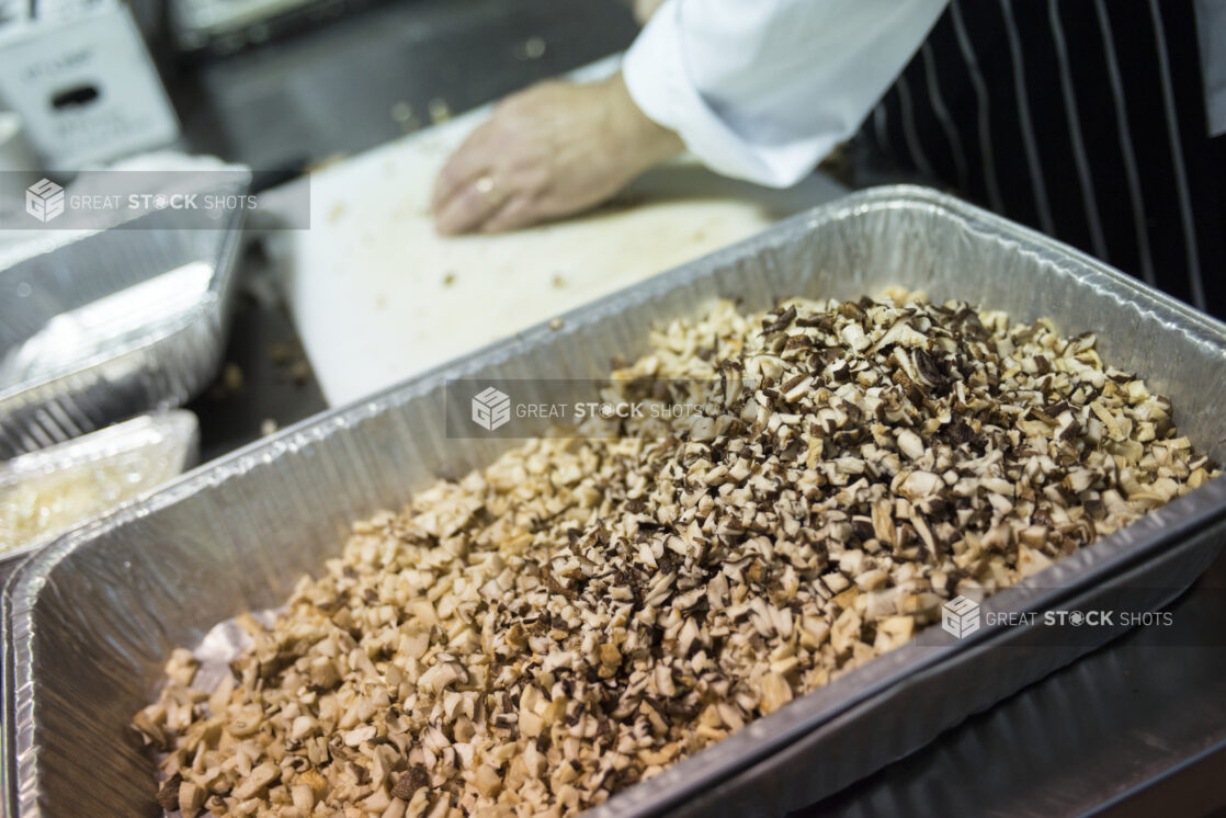 Chopped mushrooms in a full disposable hotel pan in the foreground with chef working on the cutting board in the background