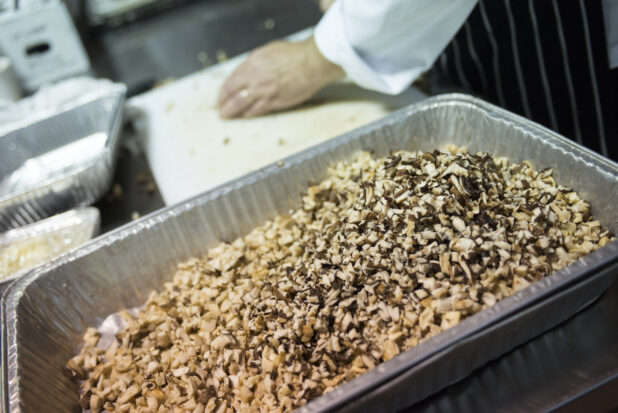 Chopped mushrooms in a full disposable hotel pan in the foreground with chef working on the cutting board in the background