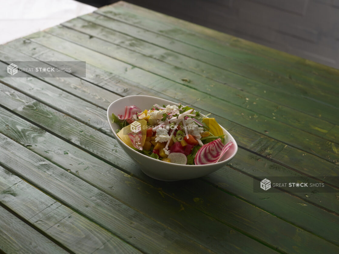 Rustic salad with greens, radishes, candy-stripe beets and goat cheese, in an angled white bowl on a green wooden table