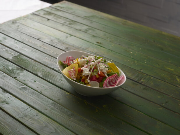 Rustic salad with greens, radishes, candy-stripe beets and goat cheese, in an angled white bowl on a green wooden table