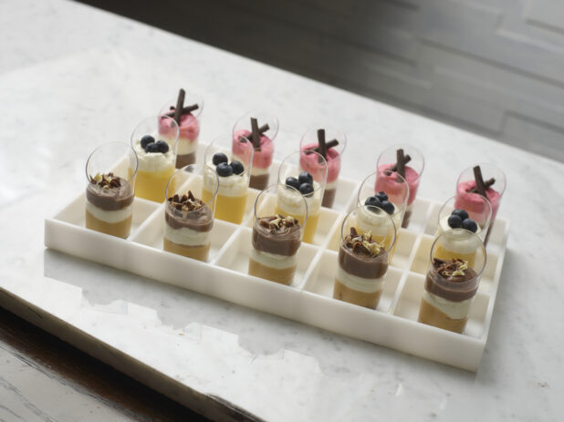 Elegant mini chocolate, mango and strawberry mousse, displayed in a white partitioned tray on a white marble background