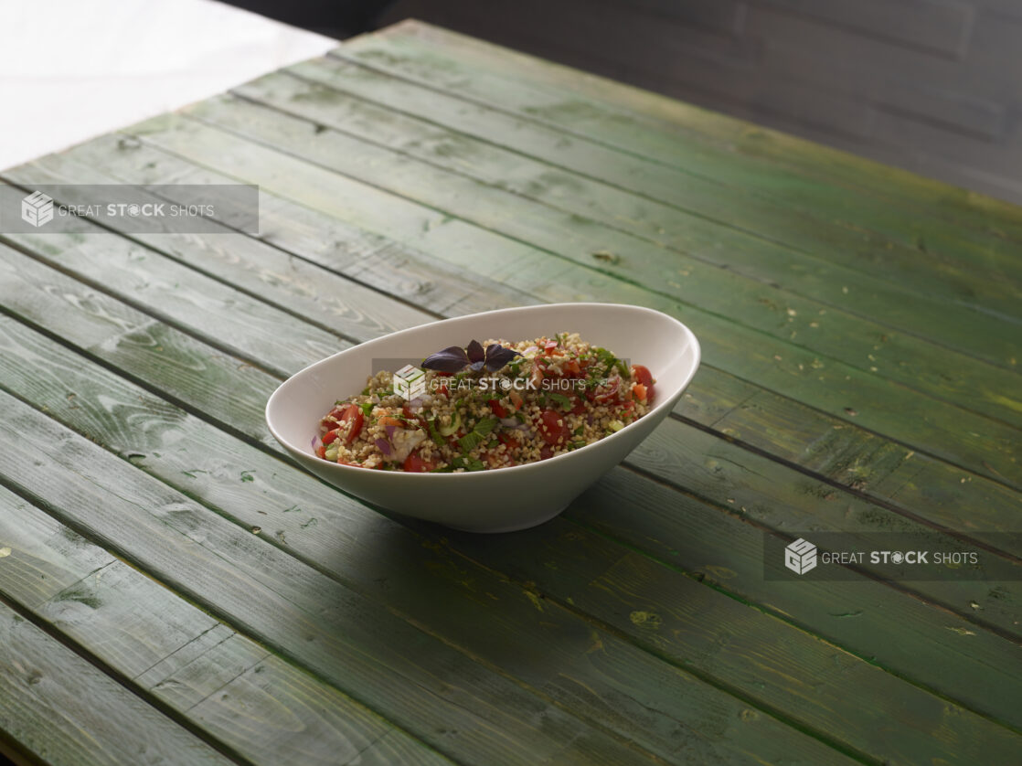 Bulgar wheat salad with fresh tomatoes, fresh basil, red bell peppers and red onion in a white bowl on a hunter green wooden table