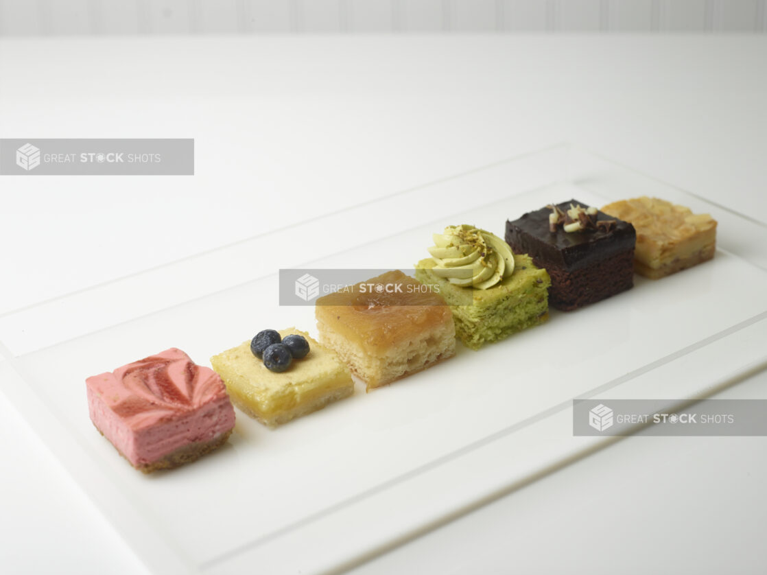 6 petit four, mini dessert cakes in a white rectangular acrylic box on a white background on a 45 degree angle