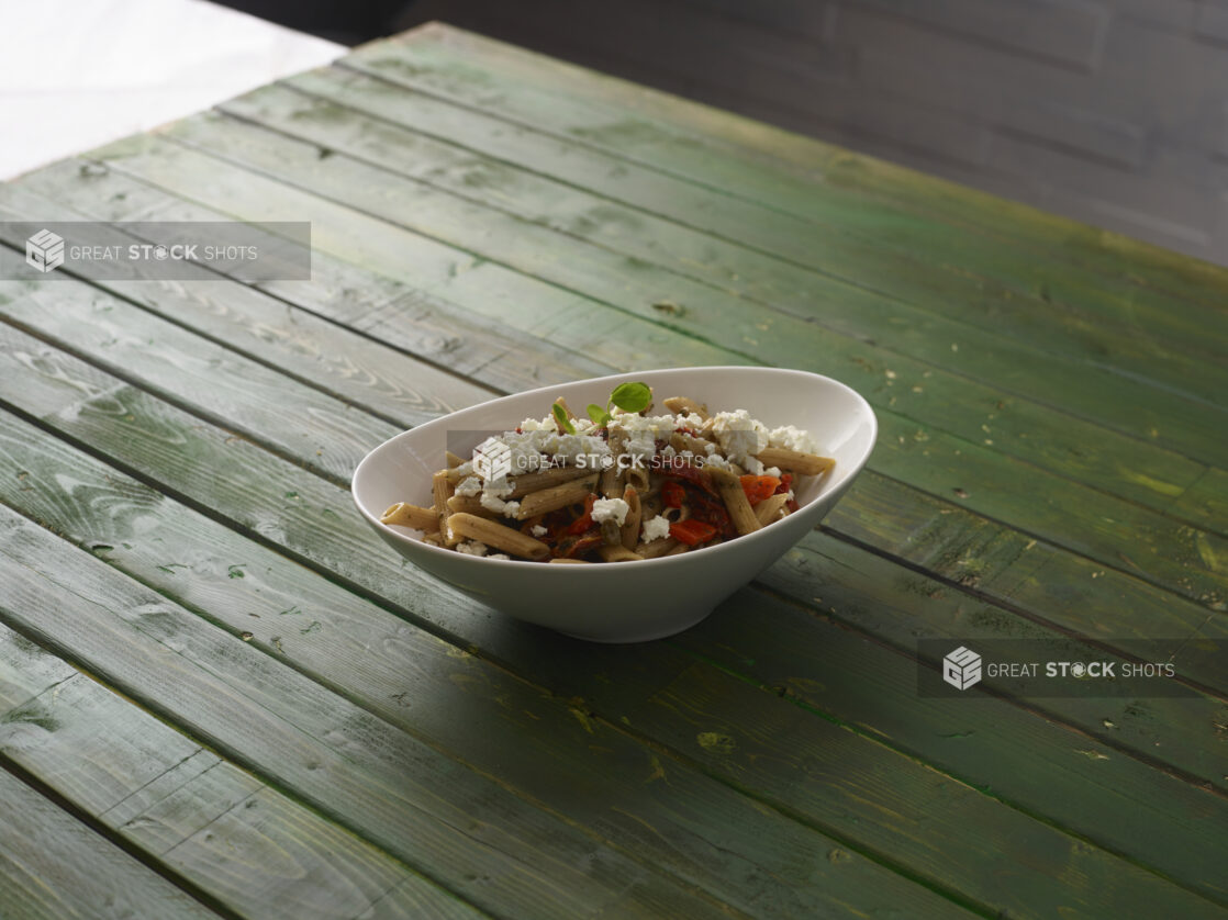 Pasta salad in an angled white bowl with penne pasta, roasted red peppers and feta cheese topped with a pea shoot garnish  on a hunter green wooden table