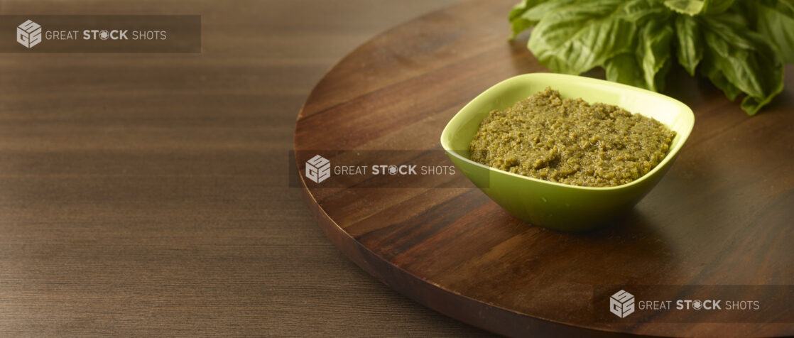 Close Up of a Square Green Bowl of Fresh Pesto Paste with a Bunch of Fresh Basil Leaves in the Background, on a Dark Wood Lazy Susan and Dark Wood Table Surface