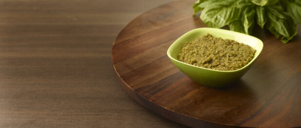 Close Up of a Square Green Bowl of Fresh Pesto Paste with a Bunch of Fresh Basil Leaves in the Background, on a Dark Wood Lazy Susan and Dark Wood Table Surface