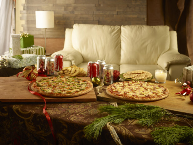 Christmas scene with 2 whole pizzas, cans of Coke and Diet Coke and pizzelle surrounded by christmas decorations
