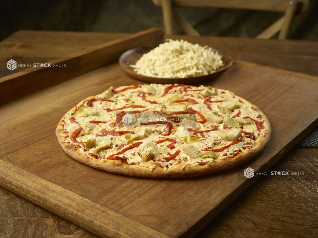 Whole vegetarian pizza with artichoke and roasted red pepper with a plate of shredded mozzarella in the background all on a wooden board