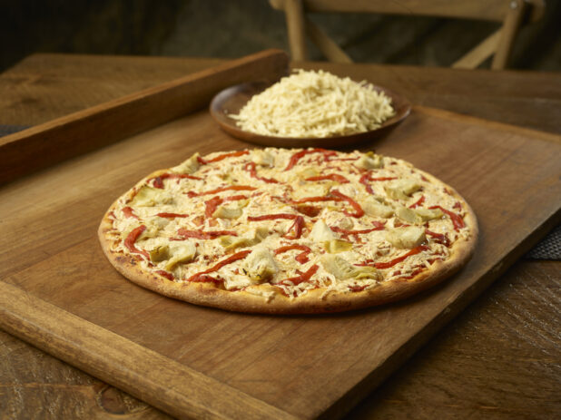 Whole vegetarian pizza with artichoke and roasted red pepper with a plate of shredded mozzarella in the background all on a wooden board