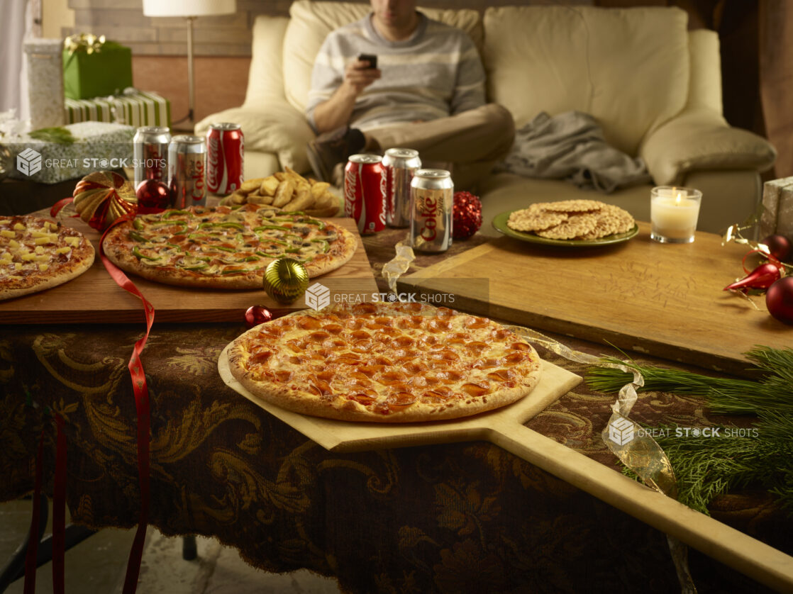 Christmas scene with 3 whole pizzas, cans of Coke and Diet Coke and pizzelle surrounded by christmas decorations with a person on a couch in the background