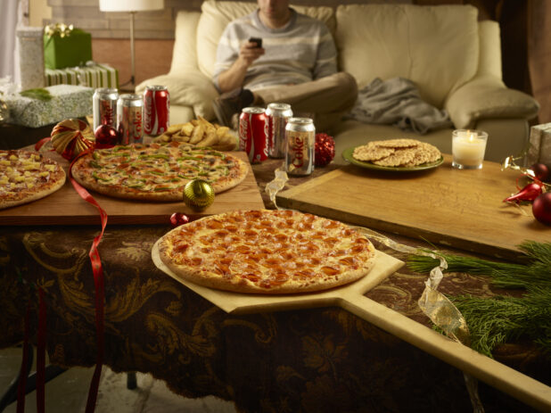 Christmas scene with 3 whole pizzas, cans of Coke and Diet Coke and pizzelle surrounded by christmas decorations with a person on a couch in the background