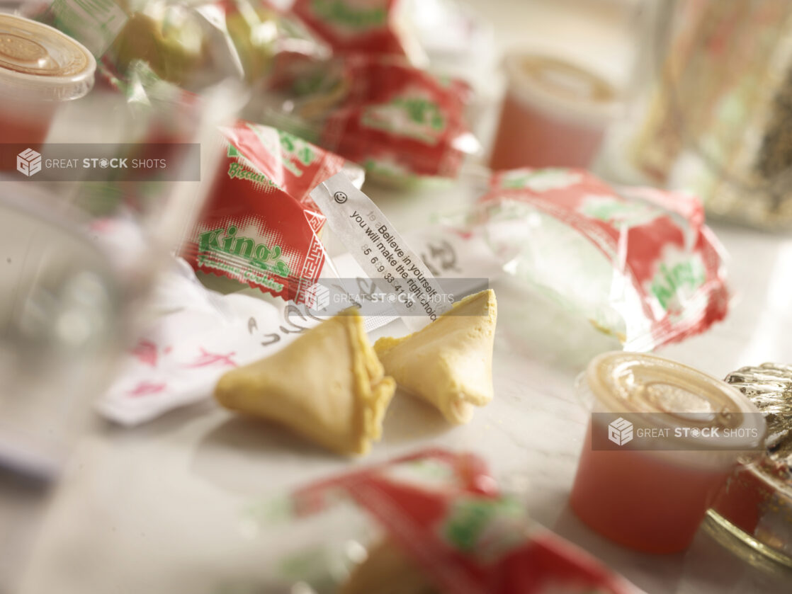 Fortune cookie broken open with fortune in foreground with cookies in wrappers surrounding, bokeh