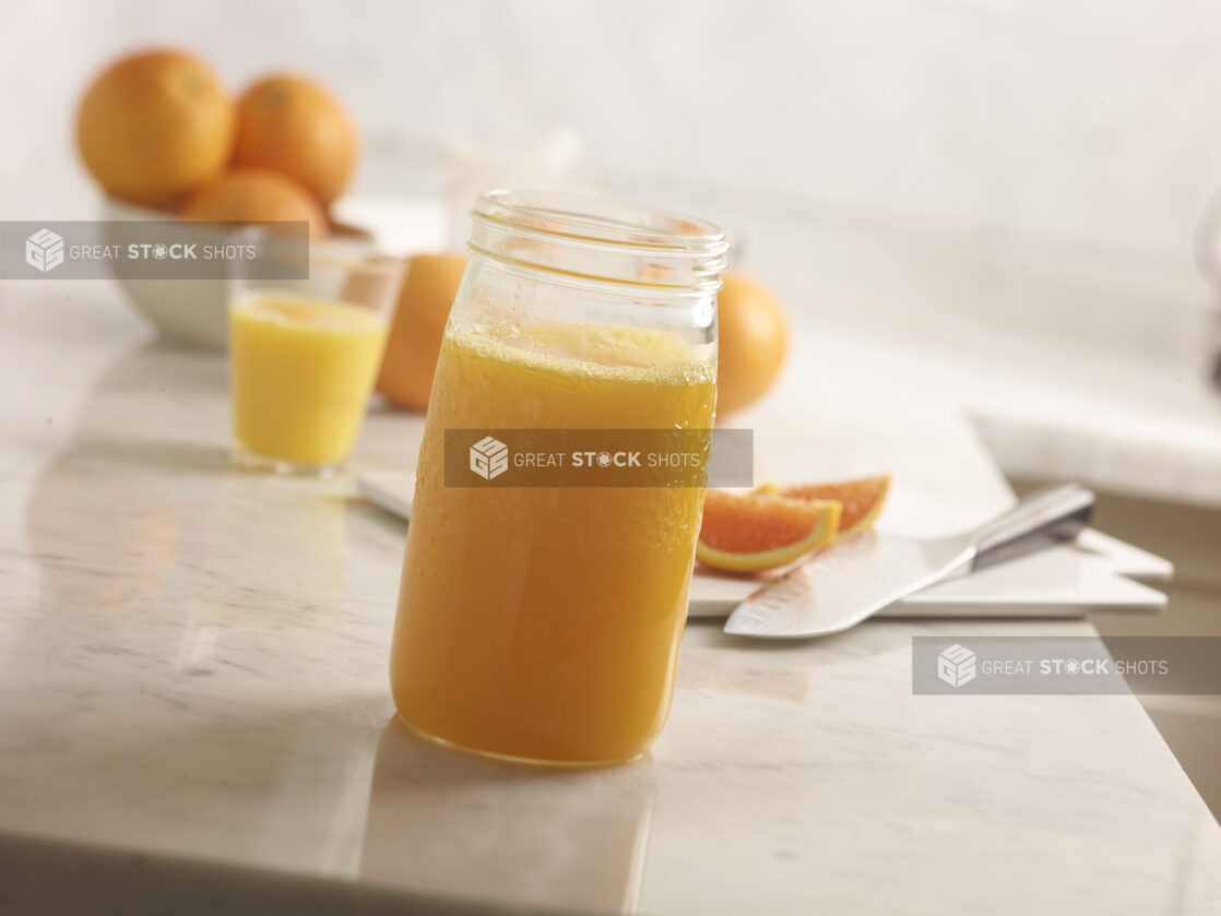 Fresh squeezed orange juice in a jar in the foreground with whole oranges, sliced oranges, cutting board and knife and glass of oj