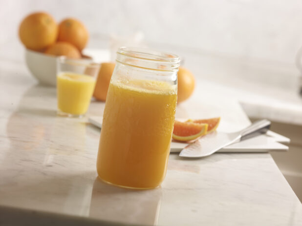 Fresh squeezed orange juice in a jar in the foreground with whole oranges, sliced oranges, cutting board and knife and glass of oj