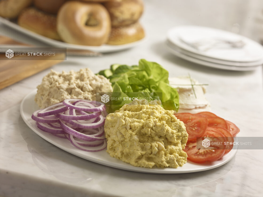 Egg salad, tuna salad, cream cheese, lettuce, sliced tomato, sliced red onion, sliced cucumber on a white round platter in the foreground with bagels in the background