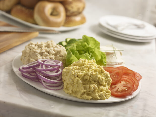 Egg salad, tuna salad, cream cheese, lettuce, sliced tomato, sliced red onion, sliced cucumber on a white round platter in the foreground with bagels in the background