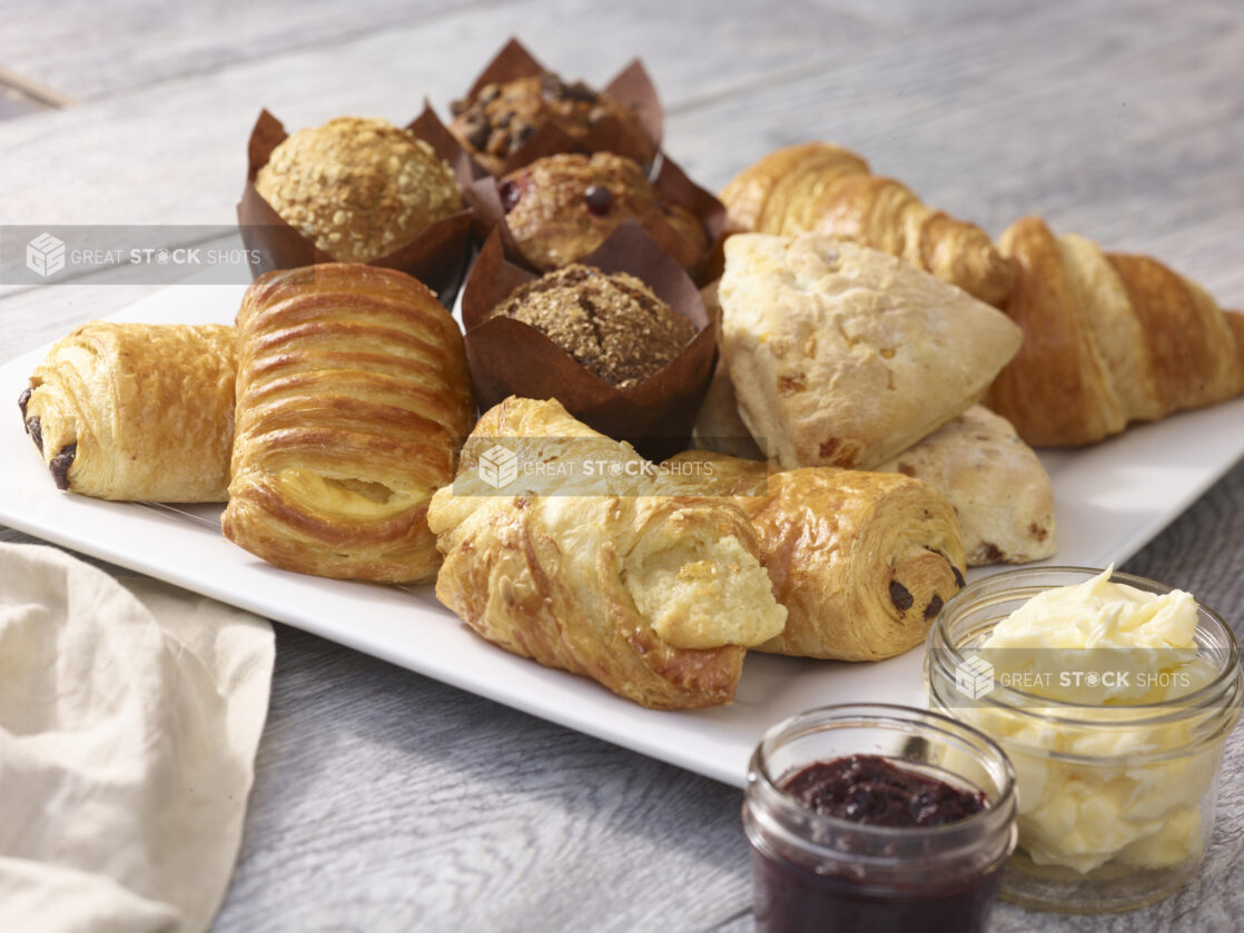 Assorted pastries on a square white platter with croissants, muffins and danish with a mason jars of butter and jam on a grey wooden background